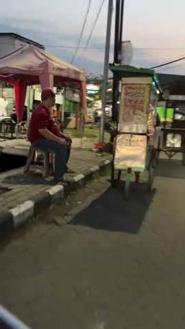 The video shows a street scene at dusk, possibly in an urban or suburban area. The first part of the video focuses on a food cart being pushed or moved. The cart appears to be colorful and has items displayed on it, possibly food. There is a person standing next to the cart, and another person in the background. The lighting suggests it is late afternoon or early evening. The second part of the video shows a three-wheeled motorized vehicle, similar to a tuk-tuk or autorickshaw, with a person riding it. The vehicle has a cargo bed at the back. The rider is wearing a green shirt. The street is paved with interlocking bricks. In the background, there are buildings, some with awnings and lights. There is also a cell tower or a similar tall structure in the distance. The sky shows colors typical of sunset or twilight. Another similar vehicle is visible further down the street. The overall atmosphere is that of a street scene with ongoing activity, likely a marketplace or a residential area with street vendors.