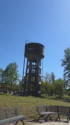 The video shows a concrete water tower in a park under a clear blue sky. The water tower has a cylindrical tank on top, supported by a framework of concrete columns and beams. A metal ladder runs up the side. The park is grassy, with trees scattered around. In the foreground, there are two metal benches. A building and a car are visible in the background. The camera is static.