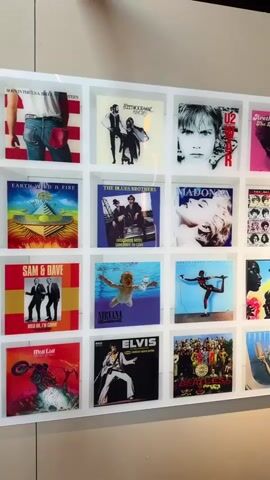 A video pans across a wall adorned with a grid of framed album covers. The display features a collection of iconic vinyl records from various artists and genres.

Starting from the left, the camera moves horizontally to the right, revealing more of the extensive collection. Prominent album covers visible include:
- Fleetwood Mac's "Rumours"
- U2's "War"
- Aretha Franklin's "The Queen of Soul"
- Earth, Wind & Fire's "Spirit"
- The Blues Brothers' "Briefcase Full of Blues"
- Madonna's "True Blue"
- Sam & Dave's "Hold On, I'm Comin'"
- Nirvana's "Nevermind"
- The Beatles' "Sgt. Pepper's Lonely Hearts Club Band" and "Yellow Submarine"
- Meat Loaf's "Bat Out of Hell"
- Elvis Presley's self-titled album
- Michael Jackson's "Thriller"
- Several covers by David Bowie, The Rolling Stones, James Brown, Supertramp, and artwork by Keith Haring.

The album covers are uniformly framed in clear, square cases and arranged neatly in a grid pattern on a light-colored wall. The setting appears to be an interior space, possibly a gallery, music store, or a decorated room.