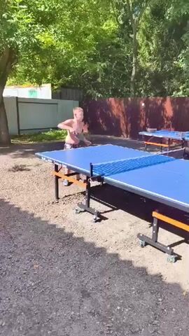 A person is playing table tennis outdoors. The person is shirtless and wearing shorts. They are on the left side of the table, facing to the right, and swinging a paddle to hit a ball. The table is blue with an orange trim and has a net in the middle. The ground is covered in gravel. In the background, there is a wooden fence and trees. There are two other table tennis tables visible in the background, slightly to the right of the main table. The lighting suggests it is daytime and sunny.