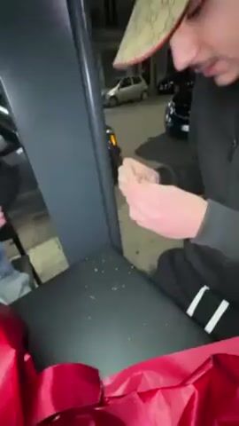 The video shows a person wearing a Gucci baseball cap and black sweat suit. They are seated outside, possibly on a black bench, with a large red ribbon and wrapping paper in front of them. The person is carefully rolling what appears to be paper or another thin material between their fingers. Another person wearing a denim vest and black beanie approaches the seated person, holding a bouquet of white and green flowers wrapped in red paper. The scene is illuminated by street lights at night, with cars parked in the background.