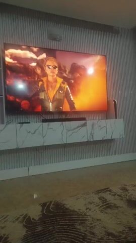 The camera films a television screen on a wall, showing a clip of the video game Mortal Kombat. A character with a green aura performs a special move. The clip then shows different characters, with a fiery background, including one wearing sunglasses and another dressed in black. The TV is mounted on a white cabinet with a marble-like pattern, set against a vertically-striped wall. A rug with a floral design is on the tiled floor.
