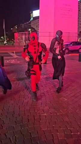 Here's a description of the video:

Three people dressed in superhero costumes are dancing in front of a large monument illuminated with pink light at night. The costumes appear to be Spiderman, Deadpool, and Venom. They are all moving in sync to music. The ground is paved with cobblestones. Cars and other people can be seen in the background, indicating they are in a public area, possibly a city square. The overall ambiance is lively and playful.