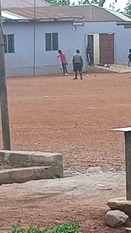 Several people are standing on a bare dirt ground in front of a one-story, light blue building with a rusty metal roof. A person in a red shirt kicks a soccer ball and another nearby person seems to be directing the action. A couple of other people are standing on the sidelines. Some concrete blocks are in the foreground, and a metal post topped with a square plate is to the right.