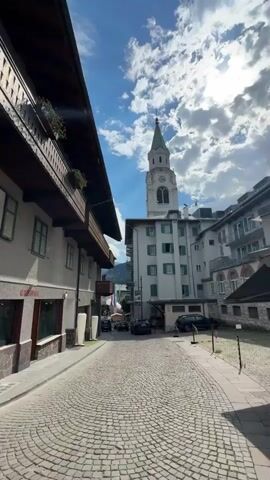 The video pans across a street in what appears to be a European town. The street is paved with cobblestones and lined with buildings. On the left, a building with wooden balconies and flower boxes is visible. In the center, a tall church tower with a green spire rises above other buildings. The buildings are a mix of architectural styles, with some featuring stone facades and arched windows. Cars are parked on the street. The sky is partly cloudy with the sun shining through, creating dramatic lighting effects. The camera moves to the right, showing a storefront with a large window displaying t-shirts. A person is reflected in the window. The scene is brightly lit, suggesting it is daytime.