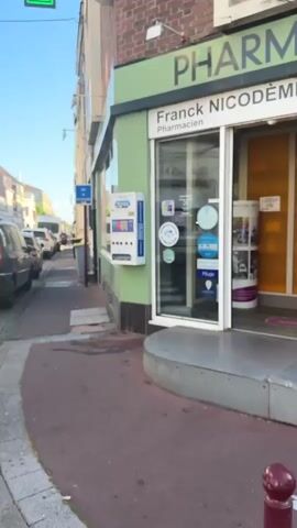The video shows a street scene in front of a pharmacy. The pharmacy has a green exterior and a glass door. On the wall next to the door is a white vending machine that dispenses Durex condoms. The vending machine has four buttons labeled 1, 2, 3, and 4, and displays images of different types of condoms. There are several cars parked on the street. A blue sign with text in French is visible on a pole. The sky is clear and the sun is shining.