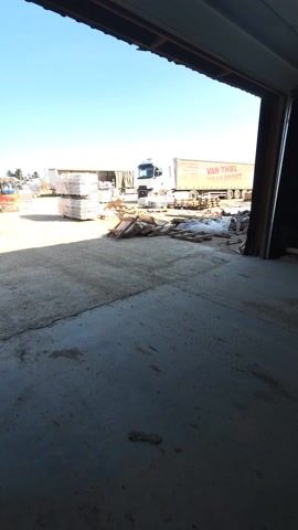 The video shows an outdoor industrial or warehouse yard, viewed from a darker, covered area, likely a loading dock or entrance. The foreground is a concrete floor that transitions into a dirt or gravel lot under bright sunlight.

In the middle ground and background, several elements are visible:
- On the right side, a large white semi-truck with a trailer is parked. The side of the trailer clearly displays red text that reads "VAN THIEL TRANSPORT".
- Further to the left, another white semi-truck trailer is partially visible.
- In the center of the yard, several tall stacks of white-wrapped pallets are present, likely containing goods or materials.
- Near the left edge of the frame, a small orange forklift can be seen moving, stirring up a small amount of dust from the ground.
- Closer to the camera, in the middle-right of the outdoor area, there are piles of wooden planks, pallets, and other debris scattered on the ground.

The sky is clear and blue, indicating good weather conditions. The bright sunlight casts distinct shadows, contrasting with the darker interior from which the video is being filmed. The overall scene suggests an active industrial or logistics operation.
