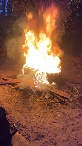 A campfire burns intensely in a dirt clearing at night. The flames are large and orange, with sparks flying upward. Wooden planks and logs surround the fire. A person's leg is visible in the bottom left corner of the frame. The camera zooms into the fire. A sound like a roaring beast is heard.