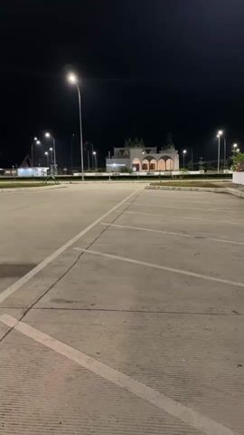 The video shows an empty parking lot at night. Streetlights illuminate the concrete surface and the surrounding area. In the background, there is a building with a dome and arched windows. A few streetlights are also visible in the distance. A black car is parked in the middle of the frame, facing away from the camera. Another white vehicle is partially visible on the left side of the frame. The ground is wet in some areas, indicating recent rain or watering. The overall scene is quiet and deserted.