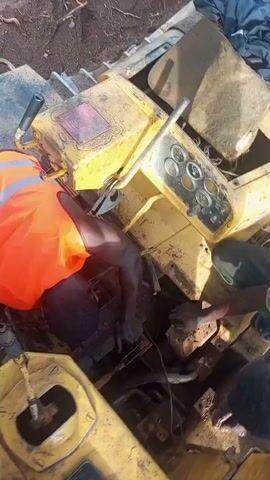 A man wearing an orange reflective safety vest is actively engaged in repairing or maintaining heavy yellow machinery. He is leaning headfirst into an open compartment of the equipment, with his hands, visibly dirty, working on internal components which include wires, hoses, and various mechanical parts. He can be seen pointing at a specific part within the machinery. Another person's hands are also visible near his, suggesting collaborative work or assistance.

The machinery features a control panel with multiple gauges and levers, indicating it is likely an excavator or similar construction vehicle. The overall color of the equipment is yellow. The scene is outdoors under bright sunlight, suggesting a clear day, possibly midday or afternoon. A smartphone rests on a yellow surface of the machinery in the foreground. The activity appears to be focused manual labor, indicating a repair or inspection task. The setting is identified as Otunade, Nigeria.
