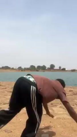 A young Black man is seen doing a handstand on a sand dune-like area near a body of water. He is wearing black athletic pants with three white stripes down the sides, a short sleeve brown shirt, and brown shoes. He stands up and smiles, then walks away from the camera. The sky is blue with some cloud cover. There are some trees and buildings visible in the background.