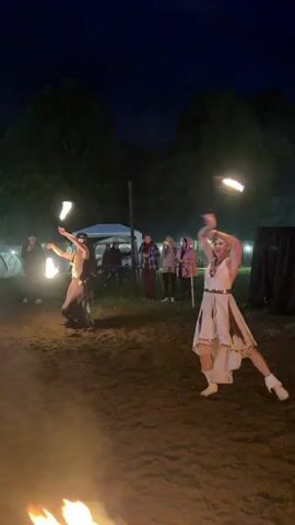 The video shows a nighttime outdoor performance. Two individuals are actively engaged in a fire-dancing routine. The person on the left, dressed in dark clothing with ornate gold accents and a headpiece, is holding flaming props in both hands, spinning them in a circular motion. The person on the right, wearing a light-colored dress and white boots, is also holding flaming props, one in each hand, and is in a dynamic pose with arms extended. Both performers are facing each other in an open area with dirt ground. In the background, a crowd of people stands watching the performance. Behind the spectators, there are tents and some artificial lighting visible, suggesting an event or festival setting. The overall atmosphere is dark, with the fire elements being the primary sources of light in the foreground.