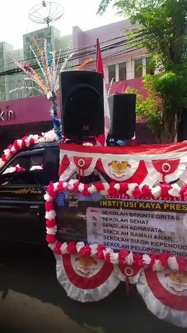 The video shows a decorated pickup truck in a parade. The truck is adorned with red and white streamers and balloons. Two large speakers are mounted on the back of the truck, along with what appears to be a flagpole with the Indonesian flag. A banner on the side of the truck displays text in Indonesian, including "INSTITUSI KAYA PRES." The background shows buildings and trees, suggesting an outdoor urban environment. The overall scene depicts a festive occasion, likely a celebration or parade.