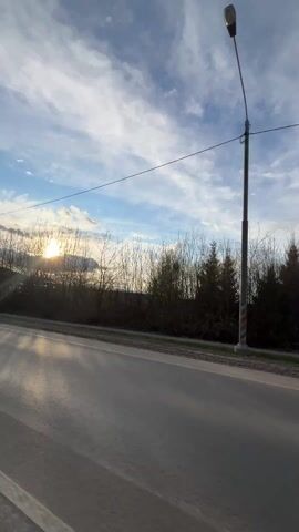 The video shows a view from a moving vehicle driving along a paved road with the sun setting in the distance. On the right side of the road is a grassy area with trees and houses. The trees are mostly bare, suggesting it may be late fall or winter. A street lamp stands on the side of the road, and power lines stretch across the sky. The sky is blue with scattered clouds. The sun's rays shine brightly, reflecting off the road's surface.