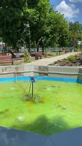 A video displays a close-up shot of a fountain with bright green water. In the background, a park is visible with trees, benches, a playground, and a paved path. The sky is blue with some clouds. The fountain is not clean, with visible debris and algae.