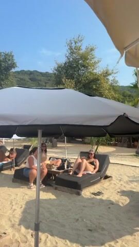 The video shows a crowded beach scene on a sunny day.  In the foreground, a white umbrella is tilted downwards, obscuring the view of the sand and several empty lounge chairs.  As the camera pans, it reveals rows of lounge chairs, many occupied by people sunbathing or relaxing.  Several large umbrellas, some white and some cream-colored, are set up to provide shade.  Palm trees and lush green trees are visible in the background, with a gently sloping hill in the distance under a clear blue sky with some scattered clouds.  People are dressed in swimwear.  Some are lying on the lounge chairs, while others are seen standing or walking along the sandy paths between the chairs.  The overall impression is a lively beach resort atmosphere.