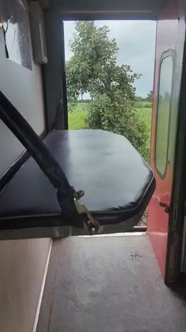 The video shows a view from inside a train compartment, looking out an open doorway. In the foreground, a folded-down seat or bench is visible, made of black, padded material with metal supports. The interior of the train appears to be functional, with some wall paneling and what looks like a handle or lever on the seat's support. Beyond the doorway, a lush green agricultural field stretches to the horizon under a cloudy sky. A few trees dot the landscape. The train tracks are visible directly outside the open door. The camera appears to be stationary, capturing the passing scenery.