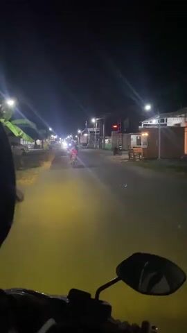The video captures a first-person perspective from a motorcycle at night. The motorcycle is moving down a paved road lined with buildings and streetlights. There are other vehicles on the road, including at least one car with bright headlights approaching the camera. Several people are visible, some on motorcycles. The scene appears to be a rural or suburban area with shops or small businesses along the roadside. The lighting is dim, with the main illumination coming from streetlights and vehicle headlights, creating a somewhat hazy atmosphere.