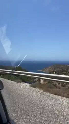 The video shows a landscape view through the side window of a moving vehicle on a sunny day. A guardrail runs along the side of the road. The road is grey and appears to be asphalt or gravel. Beyond the guardrail, there is a hillside covered with vegetation. In the distance, the view opens up to a bay with dark blue water, with landmasses on either side. The sky is a clear, bright blue.  There is a reflection on the inside of the window.