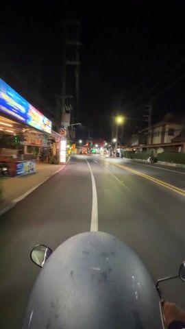The video shows a first-person perspective of someone riding a scooter down a street at night. The rider is wearing a gray helmet. The street is lit by streetlights and the lights from buildings on either side. There are a few other vehicles on the road, including cars and other scooters. The road has a white line in the middle and yellow lines on the sides. The buildings on either side of the road appear to be shops and restaurants. There is a crosswalk sign visible. The weather appears to be clear.