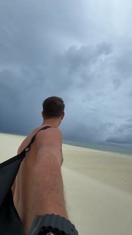 A man with short brown hair and a light complexion walks on a sandy beach, away from the camera. He is shirtless, and a black bag is slung over his shoulder. His arm is visible, and he wears a black watch with a red-and-white patterned band. The beach is flat and wide with light-colored sand that shows patterns of ripples. The ocean is in the distance. The sky is overcast with dark, ominous clouds.
