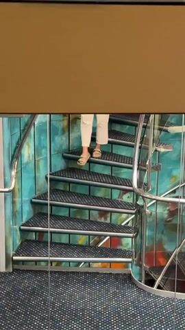 A person walks down a spiral staircase. The person is wearing white pants and a striped shirt. They are holding a phone and appear to be taking a selfie. The staircase has metallic steps with black treads. The walls surrounding the staircase are tiled with a blue and green mosaic pattern. The floor is carpeted with a dark blue and white speckled pattern. A glass railing runs along the side of the staircase. The person stops on the stairs and sits down, leaning their head on their hand while looking at the phone.