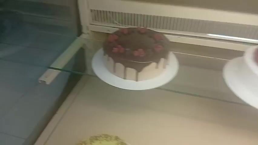 The video shows a display case of cakes. The top shelf has two cakes. One is a chocolate cake with raspberries on top. The other is a white cake with raspberries on top. The bottom shelf has three cakes. One is a light green cake topped with nuts. Another is a round cake topped with berries arranged in a circle. The third is a dark chocolate cake with a lattice pattern of white icing on top. The display case is made of glass and metal.