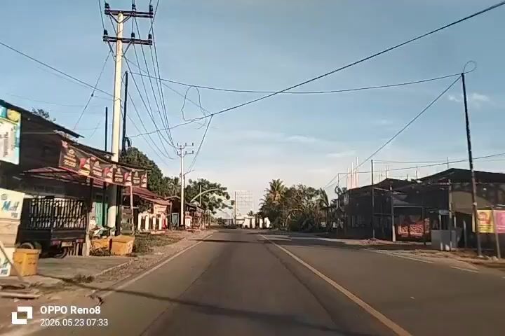 A car drives down a straight road, with buildings and trees lining either side.  Power lines run overhead. The sky is blue and mostly clear. A motorcycle appears in the distance as the car approaches an intersection. The video was captured on an OPPO Reno8, and the time stamp is 2026.05.23 07:33.