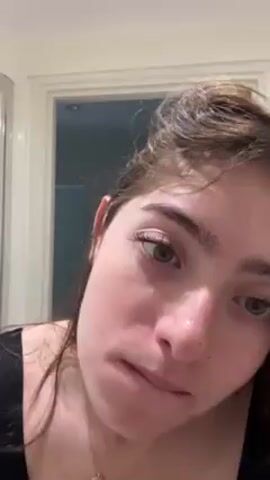 A young woman is in a bathroom, facing the camera. She appears to be applying eyebrow makeup with a small brush. The woman has light skin, light brown hair, and green eyes. She is wearing a black tank top and has a necklace on. The background shows a white door frame and a partially visible white door. The bathroom appears to be well-lit. The woman is focused on applying the makeup.