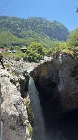 A person in a red shirt is standing at the edge of a cliff. Below the cliff, a waterfall cascades into a pool of water. The surrounding environment is a mountainous area with trees and a river.