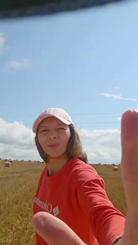A woman wearing a pink baseball cap and a red long-sleeved shirt runs through a field of hay bales under a blue sky with scattered clouds. The woman starts running towards the right of the frame, then jumps in the air.