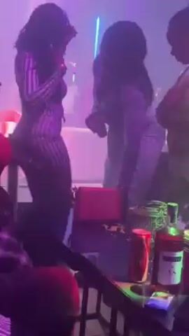 The video shows two women dancing in a crowded nightclub. Both women have long, dark hair. The woman in the foreground is wearing a fitted, vertically striped dress and holding a red purse on her shoulder. The other woman appears to be wearing a light-colored, fitted dress. The lighting in the club is dark with colored lights flashing across the scene. Other people are visible around the table, and there are bottles and cans on the table.