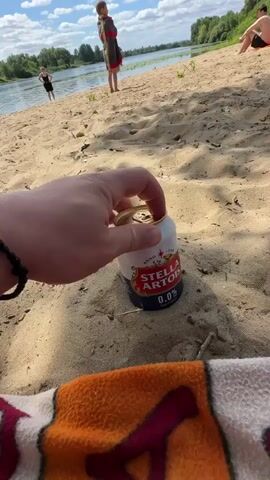 A hand opens a can of Stella Artois 0.0% beer on a sandy beach. In the background, a river is visible, with trees on the opposite bank. Two children and one adult are in the water, and another adult is sitting on the sand.