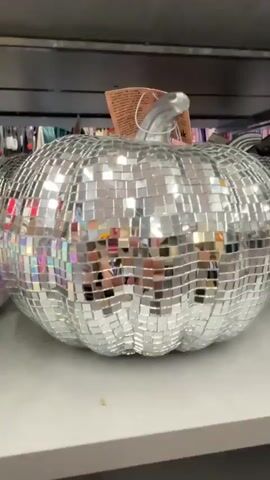This video pans across a display of disco ball pumpkins. The pumpkins are round with the typical pumpkin ridges and have a silver stem. They are covered in small, square, mirrored tiles that reflect the light, giving them a sparkly appearance. The pumpkins are arranged on a shelf, likely in a retail store, with other decorative items and clothing visible in the background. The lighting is bright, enhancing the reflective quality of the pumpkins. The camera moves slowly, allowing viewers to see the details of the pumpkins from different angles.