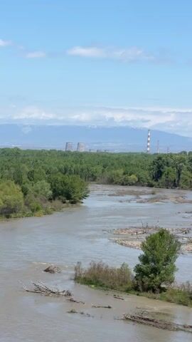 The video shows a wide river flowing through a lush, green landscape. In the background, a power plant with large cooling towers can be seen, set against a backdrop of distant mountains with some snow. The sky is blue with scattered white clouds. The river is wide and murky, with some visible debris and small islands of vegetation. The camera slowly zooms in on the power plant and cooling towers, highlighting their industrial presence in the natural setting.