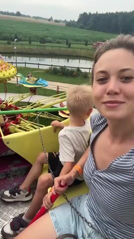 The video shows a woman and a child on a carnival ride. The woman is in the foreground, smiling at the camera and holding onto a red bar. The child is seated behind her. The ride appears to be a spinning amusement park attraction with yellow and red elements. In the background, there is a green landscape with trees and a pond. Another carnival ride with blue boats is visible in the distance. The sky is overcast.