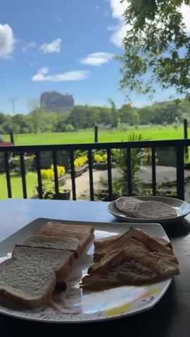 A static, eye-level shot presents a partially obscured view of Sigiriya Rock in Sri Lanka, framed by a breakfast table and a balcony railing. The table displays a plate of untoasted and toasted bread, alongside another plate with round, flatbread. The scene is set outdoors, with a view of a lush green field leading to Sigiriya Rock, under a bright blue sky with scattered clouds. The foreground includes sections of a black balcony railing and potted plants, contributing to the relaxed outdoor dining setting.