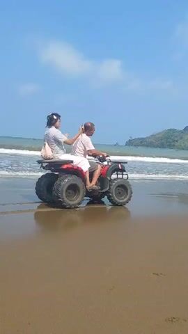 The video shows two people riding an ATV on a sandy beach. The person in front is driving, and the person behind is holding up a selfie stick and taking a photo or video. The ATV is red and black, and the people are wearing casual clothing. The beach is wide and sandy, with waves gently rolling in from the ocean. In the background, there are green hills covered in vegetation, and the sky is blue with some scattered clouds. The overall scene depicts a leisurely day at the beach.