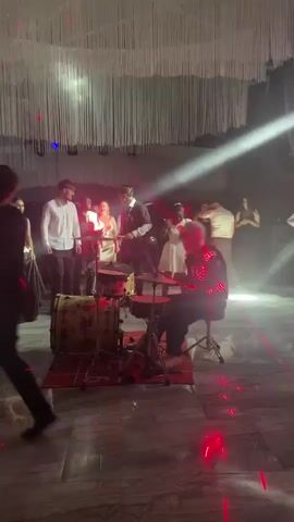 The video shows a group of people in a dimly lit room with purple and red stage lights. In the foreground, a person with dark hair is walking away from the camera. To the right, a person wearing a black outfit with illuminated red accents is seated at a drum set and appears to be playing. In the center of the frame, another person in a black top hat is standing and holding drumsticks, also appearing to be playing drums. Other individuals are visible in the background, some appearing to be dancing or watching the performance. The environment suggests an indoor event, possibly a party or concert, with a stage or performance area set up with musical instruments and lighting.