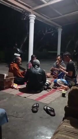 Five adult males are seated on the floor around a woven mat. They are in an outdoor covered area, likely a porch or carport, at night. The ground is concrete. The men appear to be engaged in an activity, possibly playing cards or a board game, as they are looking down and interacting with items on the mat. One man on the far left is wearing an orange hooded sweatshirt. Another man in the center is wearing a dark leather jacket and a cap. A box and some food items are on the mat. Two pairs of flip-flops are visible on the concrete in the foreground. The background is dark and suggests vegetation and trees. Two motorcycles are parked behind the group. The lighting is artificial and casts shadows.
