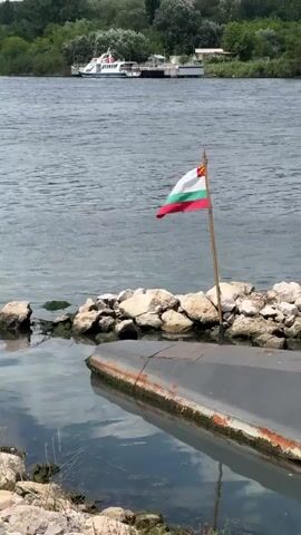 The video begins with a close-up of the Bulgarian flag on a pole in the water. The camera then pans right, showing the water and some rocks on the shore. A large, dark object, likely a submerged boat or submarine, is partially visible. The camera continues to pan right, revealing more of the dark object, which appears to be a submarine, with a ramp leading up to it. The scene then expands to show the submarine docked by the shore, with a body of water and trees in the background. A passenger ferry is visible on the water in the distance. The video ends by showing the side of the submarine, the water, and the shore with trees and buildings in the background under a cloudy sky.