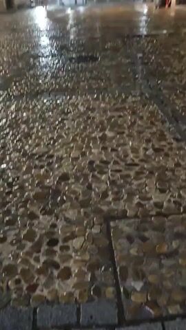The video begins with a close-up of wet, cobblestone pavement at night. As the camera pans up, it reveals a town square at night with buildings on either side. The pavement is glistening from rain, and there are streaks of rain visible in the dark sky. A few people are seen standing or sitting outside the buildings. In the foreground, the cobblestones are clearly visible, reflecting the light from nearby lamps. The overall atmosphere is dark and wet, suggesting it is raining or has recently rained.