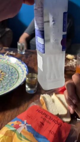 A group of men are sitting at a wooden table. In the foreground, a bottle of vodka is being held over the table, with the liquid pouring into a small shot glass. Slices of white bread are visible on the table, as is a colorful patterned plate. Other shot glasses and an orange drink are also present. One man in the background is wearing a dark t-shirt, and another is wearing a blue t-shirt. The setting appears to be indoors with blue walls in the background.