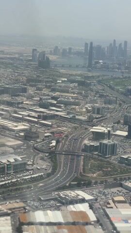 The video shows an aerial view of Dubai, United Arab Emirates.  The camera is likely positioned inside an airplane as part of the wing is visible. In the background, a cluster of skyscrapers are seen. A bridge spans over a body of water. The foreground is dense with buildings, roads, and highways. Traffic is visible on the roads. The sky is hazy.