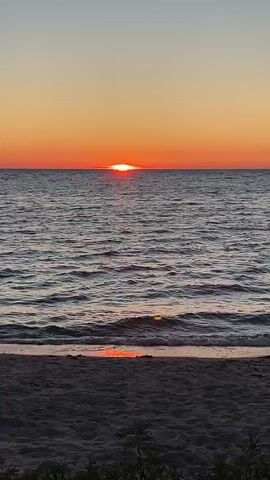 The video shows a sunset over a body of water. The sun is a bright orange disk just above the horizon, casting a warm glow on the sky. The water is calm with gentle waves lapping at the shore. The beach in the foreground is sandy with some green foliage at the bottom edge of the frame. The colors in the sky range from a pale yellow near the horizon to a soft orange higher up. The water reflects the colors of the sunset, creating a shimmering effect. There are no people or man-made objects visible in the scene.