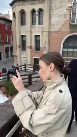 A person in a beige trench coat takes a picture with a black camera while standing on a bridge with a wooden railing. The view pans to the right, revealing a canal with various buildings, boats, and people on the sidewalks along the canal. The sky is overcast with clouds.