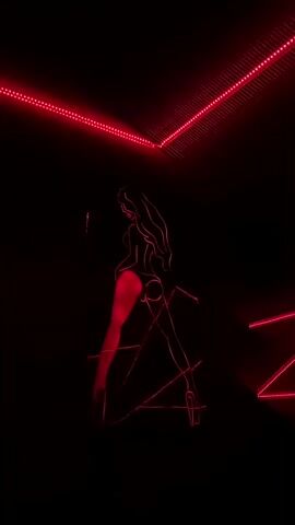 A neon outline of a woman in a provocative pose is illuminated against a black background. Red neon lights trace the figure, with the legs and buttocks highlighted in a brighter red. The figure appears to be wearing high heels. Red neon strips are also visible along the ceiling and a wall, forming a geometric pattern.