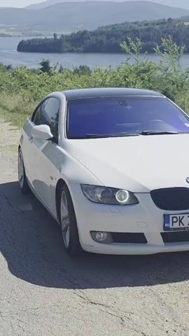 A white BMW coupe with a black roof and tinted windows is parked on a paved road. The car is positioned at an angle, facing forward and slightly to the right, with its front end visible. Behind the car, there is a body of water, likely a lake, bordered by lush green trees and rolling hills in the distance under a clear, sunny sky. The road appears to be slightly cracked and worn. No people are visible in the video. The license plate is partially visible and reads "PK 7359".