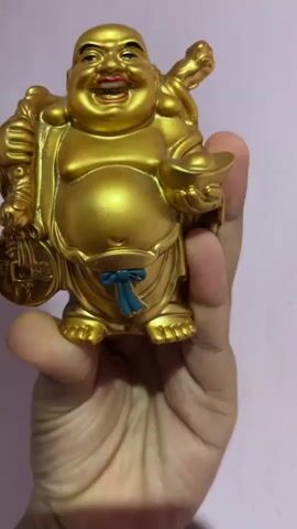 A close-up, eye-level shot shows a hand holding a golden statue of a laughing Buddha. The statue is detailed, with a round belly, a smiling face, and adorned with a blue sash. The Buddha holds a coin in one hand and a gold ingot in the other. The background is a soft, pinkish-purple. The lighting is soft and warm, illuminating the gold of the statue.