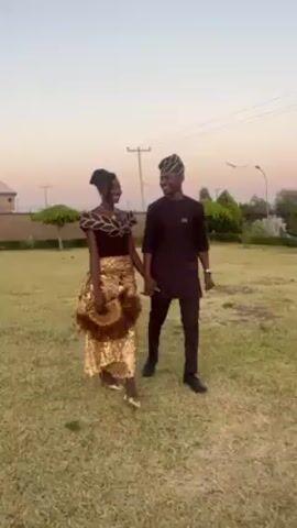 A young adult couple, a woman and a man, stroll hand-in-hand across a grassy lawn under a clear, slightly purple-tinged sky. The woman, who has dark skin, wears a brown, fitted top with decorative dark trim and a gold, sequined, fringed skirt. She carries a matching fan. The man has dark skin and short hair, and he wears a dark brown tunic-style shirt and matching pants with a patterned hat. They are both smiling and looking at each other. In the background, there are buildings, utility poles, decorative shrubs and lampposts.